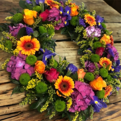 Bright Floral Wreath - Gerberas, hydrangea, iris, roses, seasonal flowers and greenery. It is arranged on a wet floral foam frame. Easy to carry. Different colours can be requested in the notes. Perfect for a funeral tribute, memorial or grave.