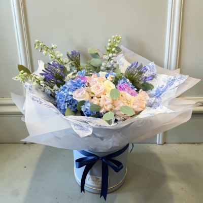 The Bridgerton - Inspired by timeless romance and refined elegance, The Bridgerton is a bouquet worthy of the most graceful leading lady — your mum.

This exquisite arrangement blends soft blush and cream roses with delicate hydrangeas in dreamy shades of powder blue and pastel pink. Lavender-toned blooms and sculptural purple accents add a regal touch, while fresh eucalyptus and seasonal white florals bring lightness and texture.

Hand-tied and presented in a luxury powder-blue hat box, finished with a deep navy satin ribbon and layered toile wrapping, this design captures classic English garden charm with a modern, couture finish.